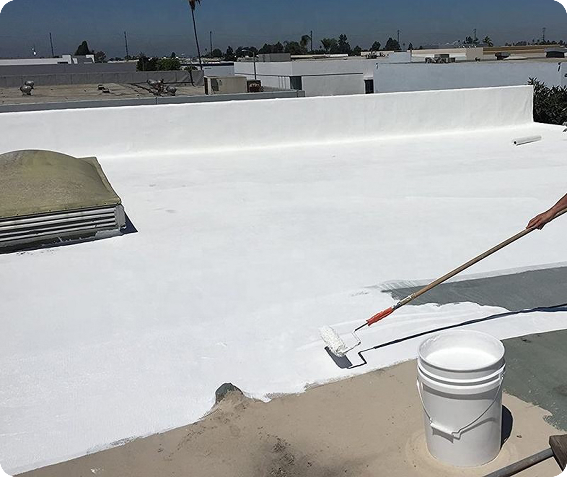 Person painting a white rooftop with a long-handled roller beside a bucket under a clear sky