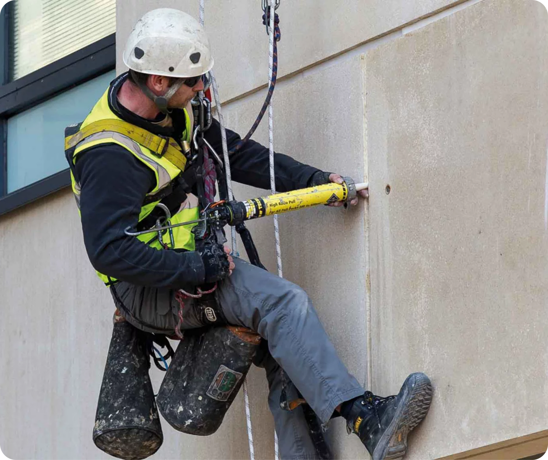 Worker in safety gear rappels down a building wall, using a yellow drill or tool near a seam.