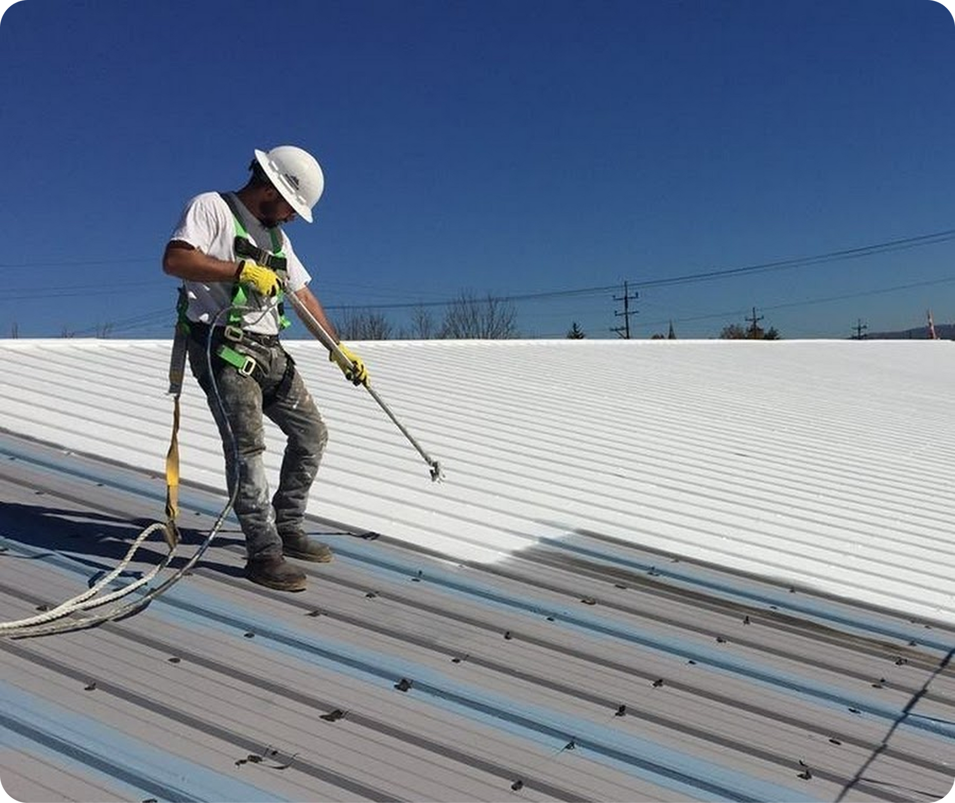 Worker in a hard hat spray-coating a metal roof under a clear blue sky