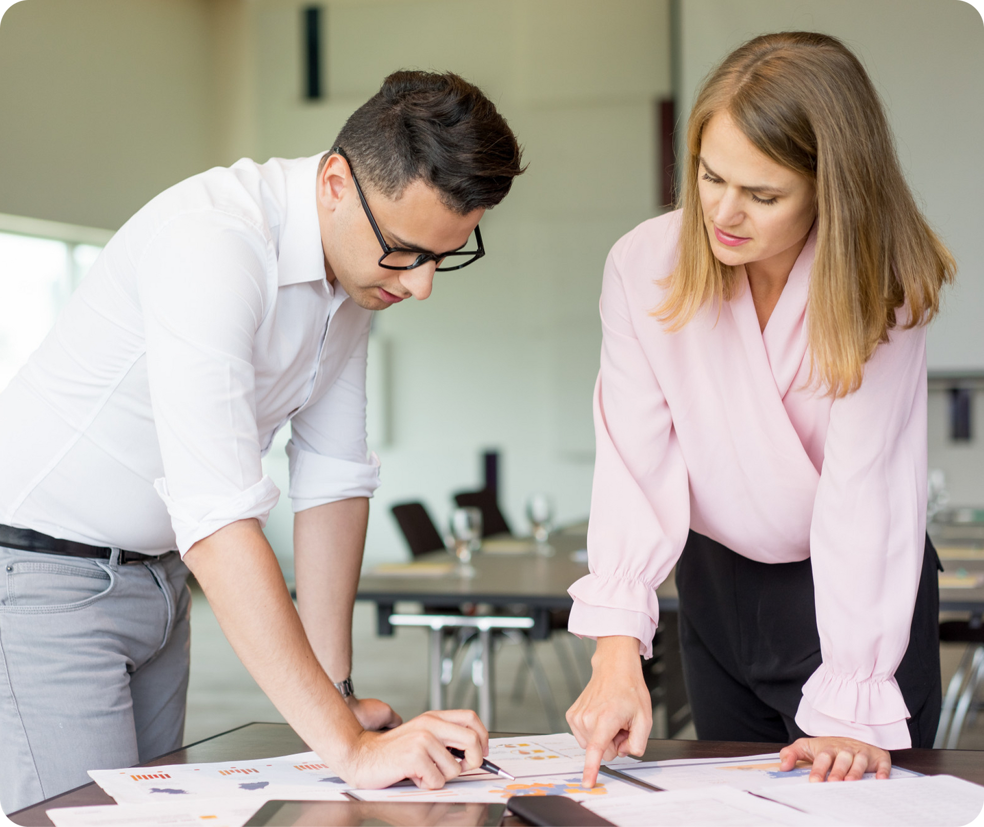 Two coworkers lean over papers on a table in a bright office, discussing a project.