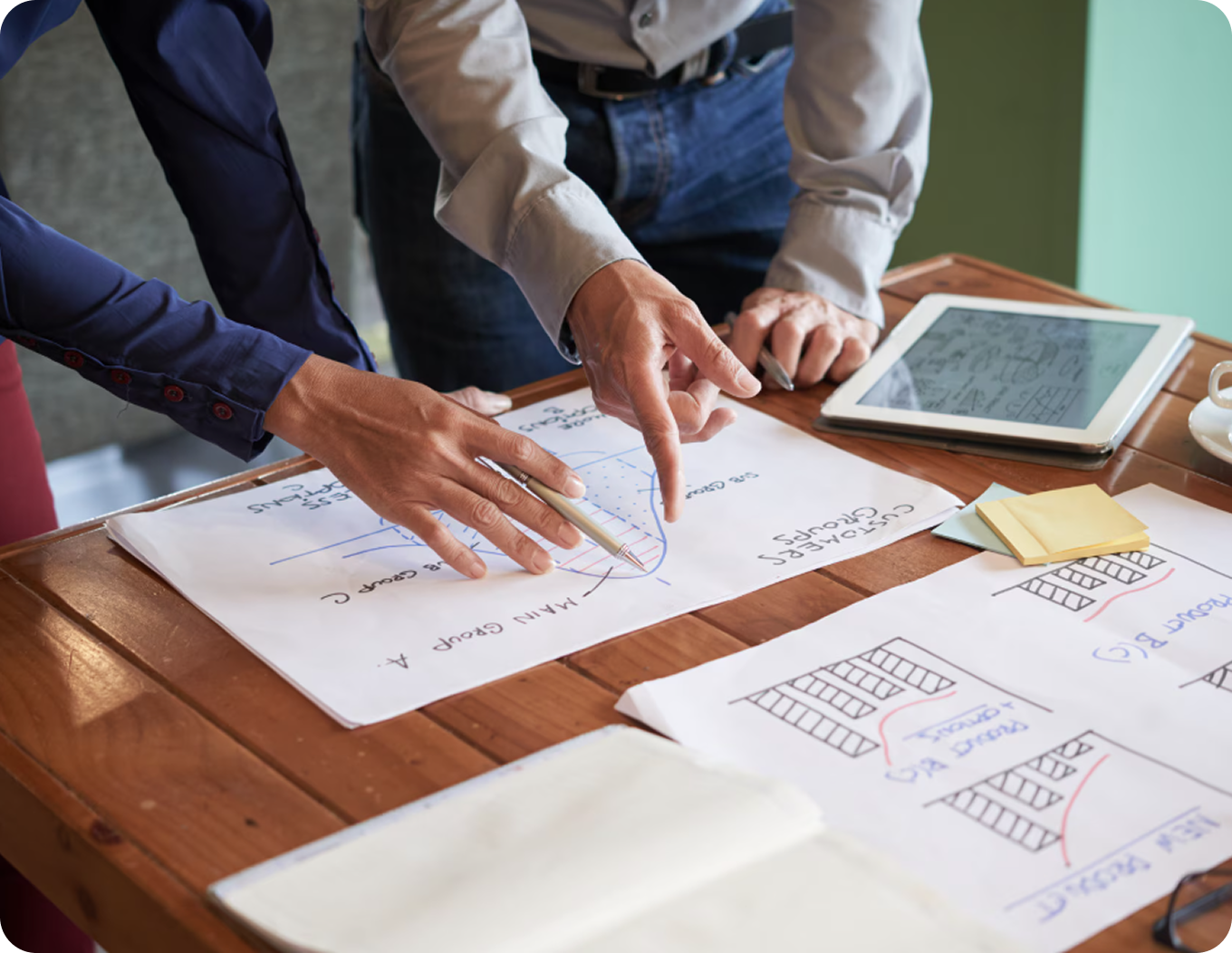 Hands pointing at house sketches and floor plans on a table with a tablet nearby