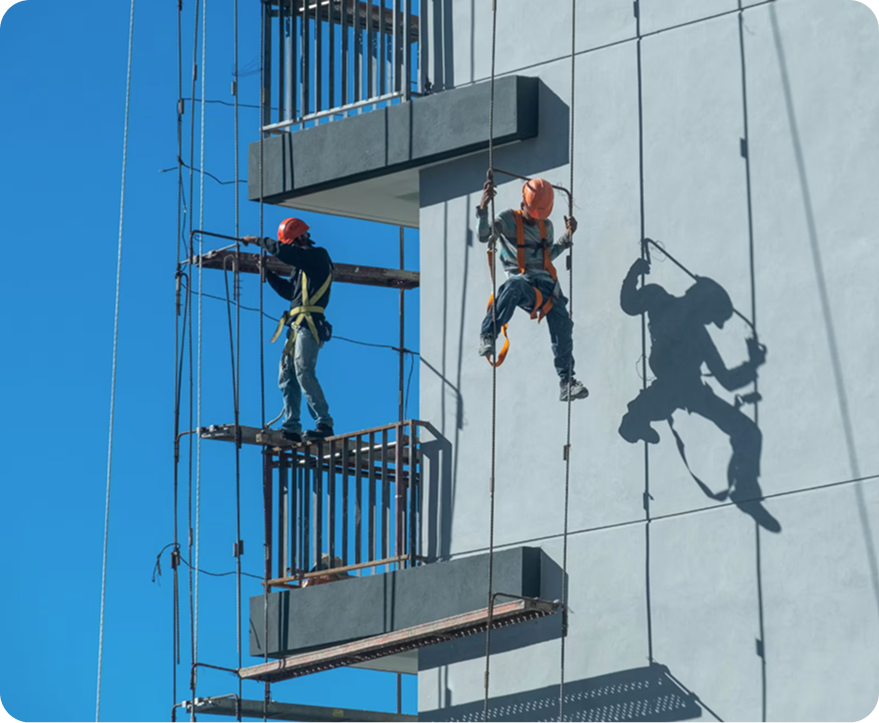 Two window cleaners on ropes beside a tall blue building, one in red helmet and one in orange gear.