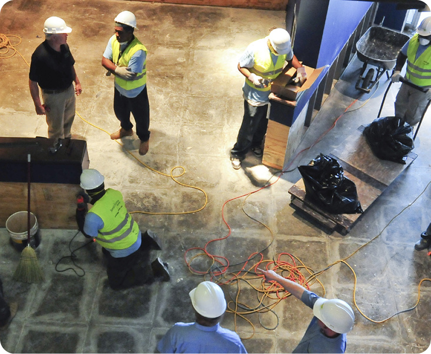 Construction workers in white hard hats inspecting a concrete floor with cables and tools indoors