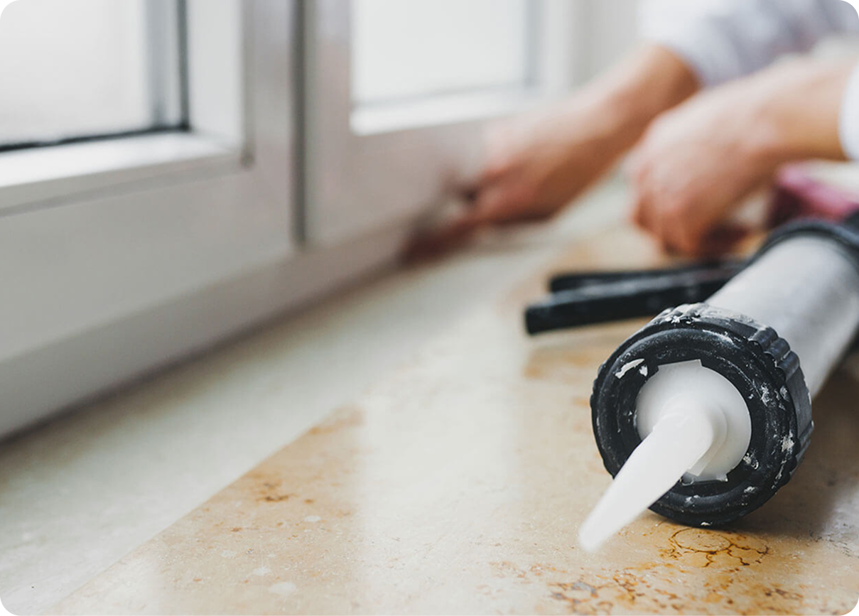 Caulking gun applying white sealant along a window frame beside a hand