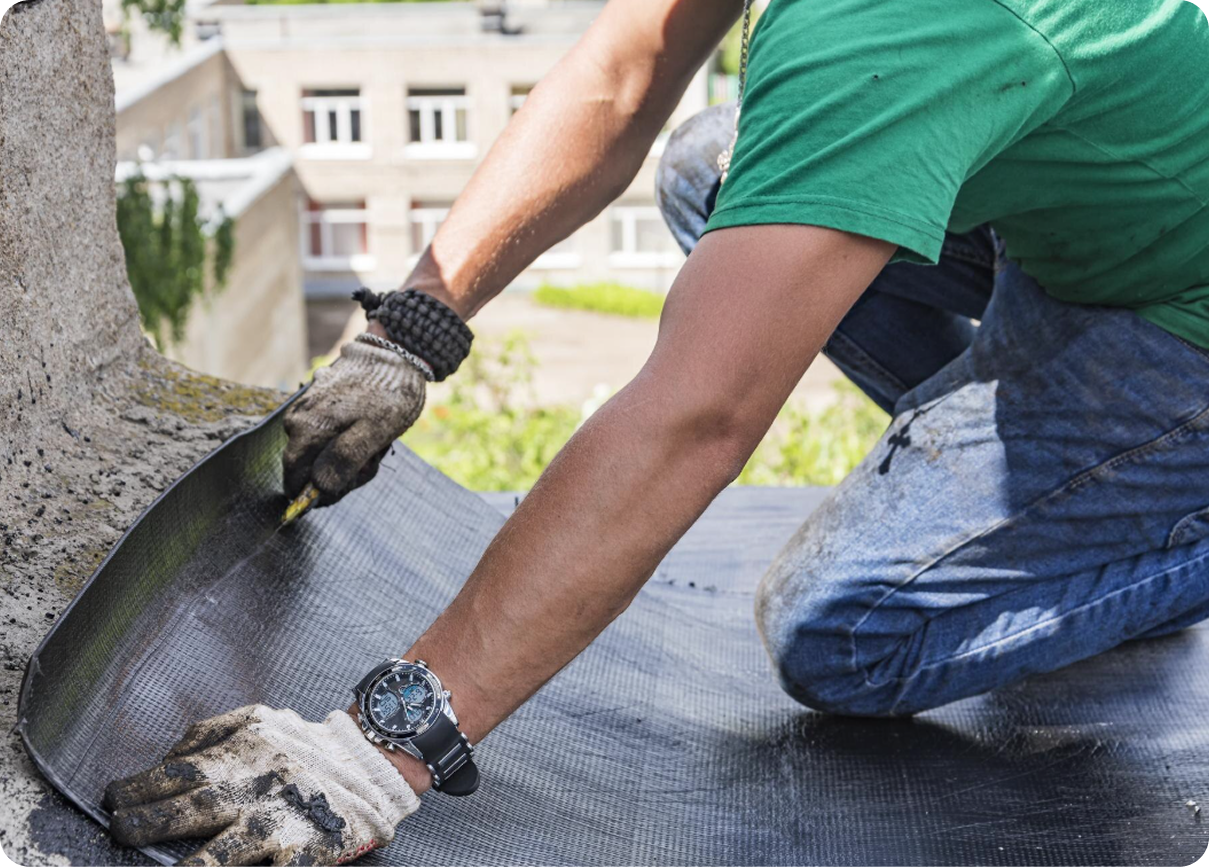 Gloved hands smoothing black roofing material on a rooftop near a tree trunk