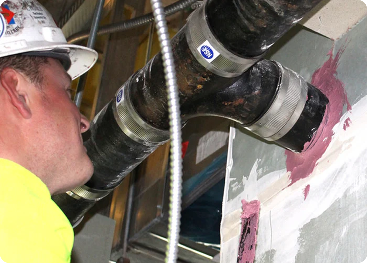 Worker in a hard hat inspecting large black pipes in a construction wall cavity