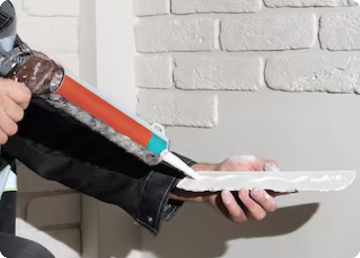 Person using a caulking gun to apply white sealant along a surface against a brick wall.