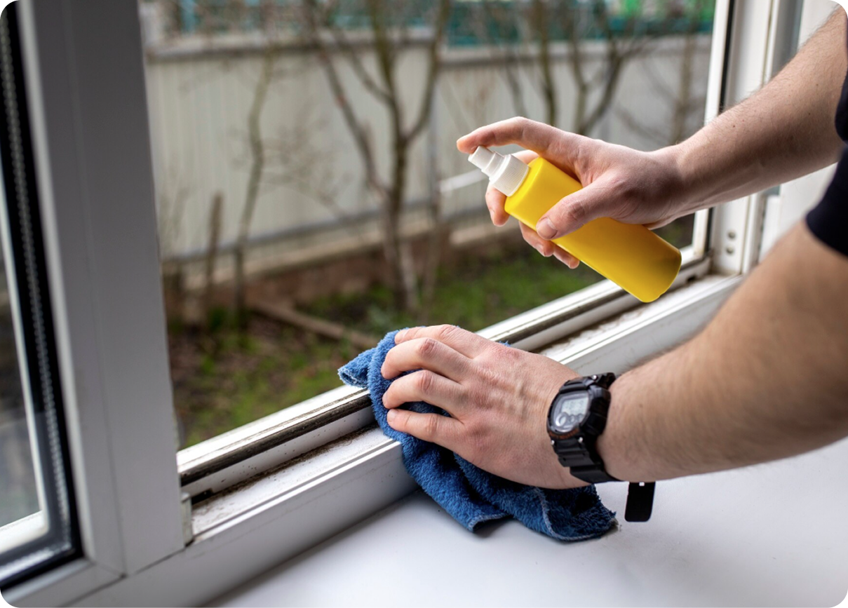 Hands cleaning a window sill with spray bottle and blue cloth
