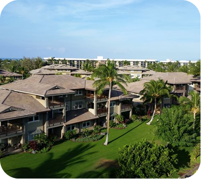Aerial view of a tropical resort with tiled-roof buildings, palm trees, and green lawns under a blue sky
