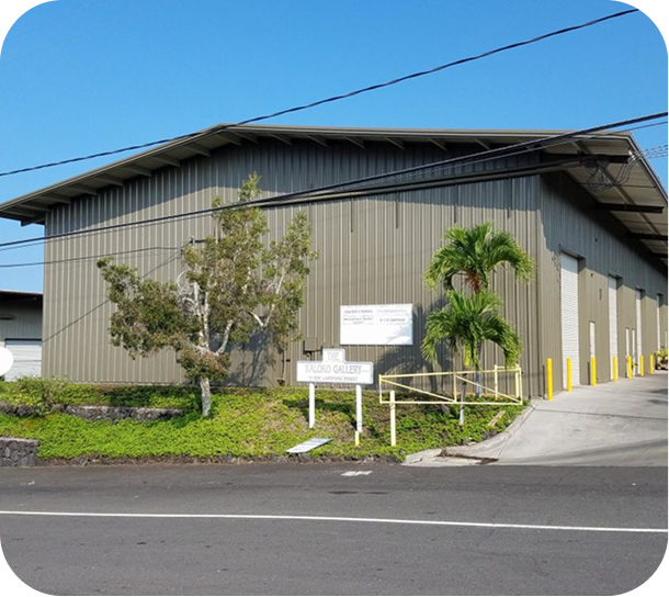 Gray industrial warehouse beside a road, with a small sign, palm tree, and yellow bollards under a blue sky