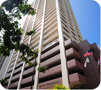 Tall beige hotel tower with rows of balconies, seen from below beside trees and a U.S. flag.
