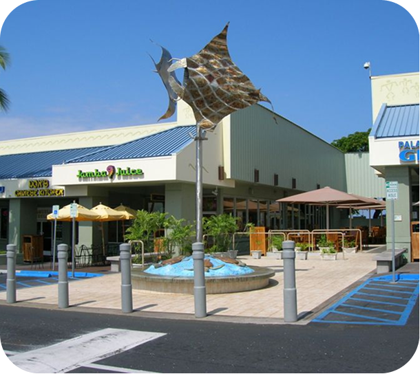 Outdoor shopping center entrance with umbrellas, fountain, and a large fish sculpture over the roof