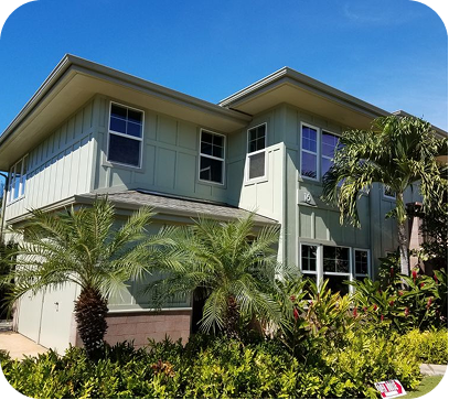 Two-story pale green house with palm trees and lush tropical landscaping under a bright blue sky