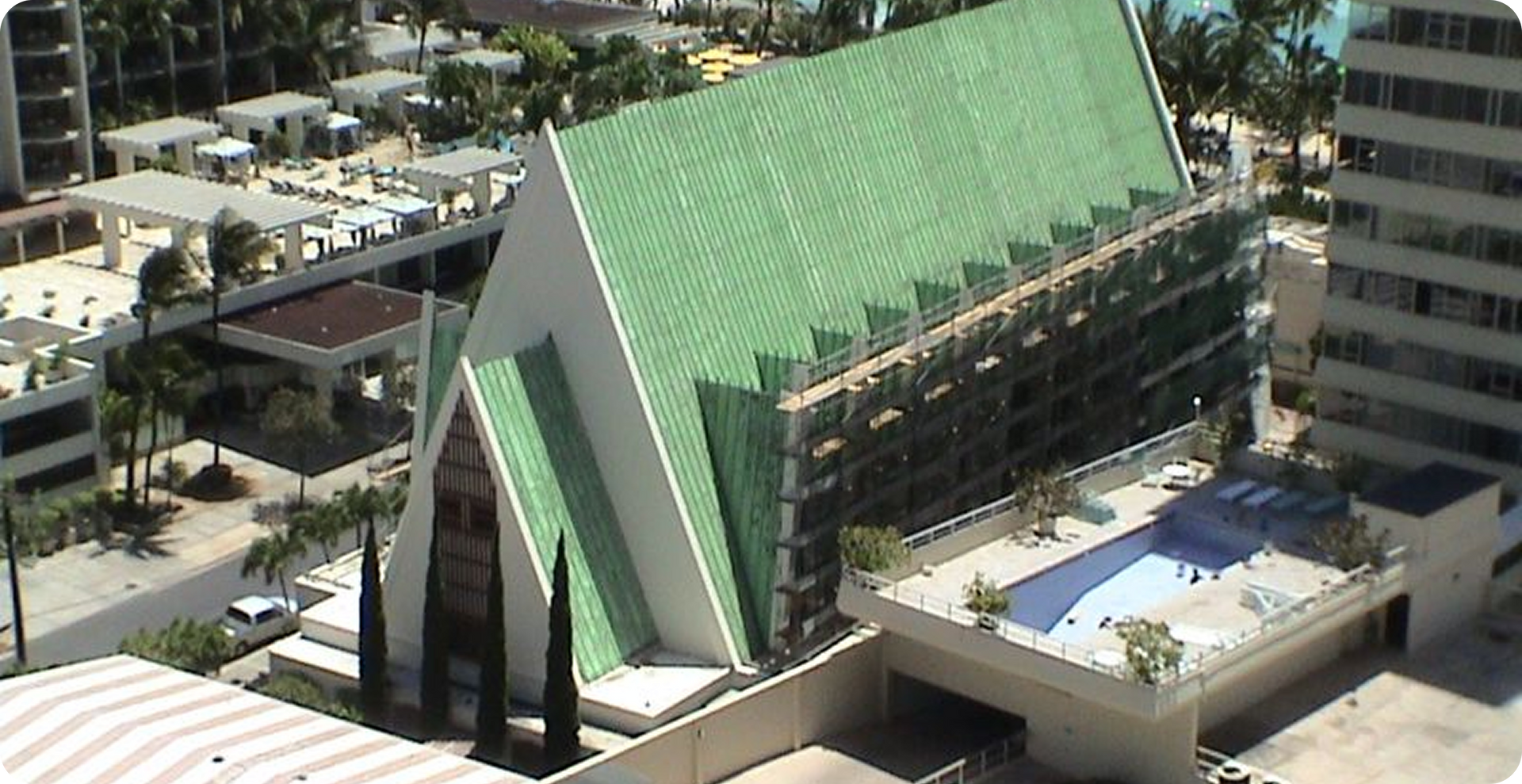A green-roofed A-frame building in a city, viewed from above, with surrounding rooftops and trees.