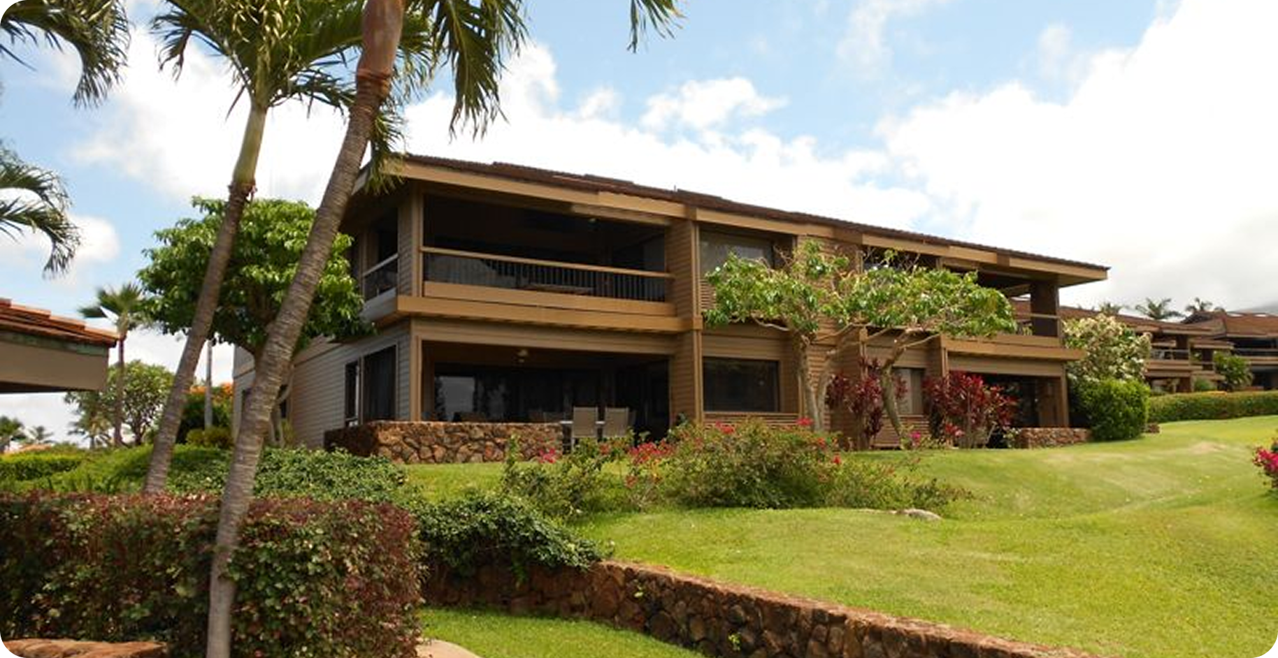Modern two-story resort building with balconies, surrounded by a green lawn and palm trees under a blue sky