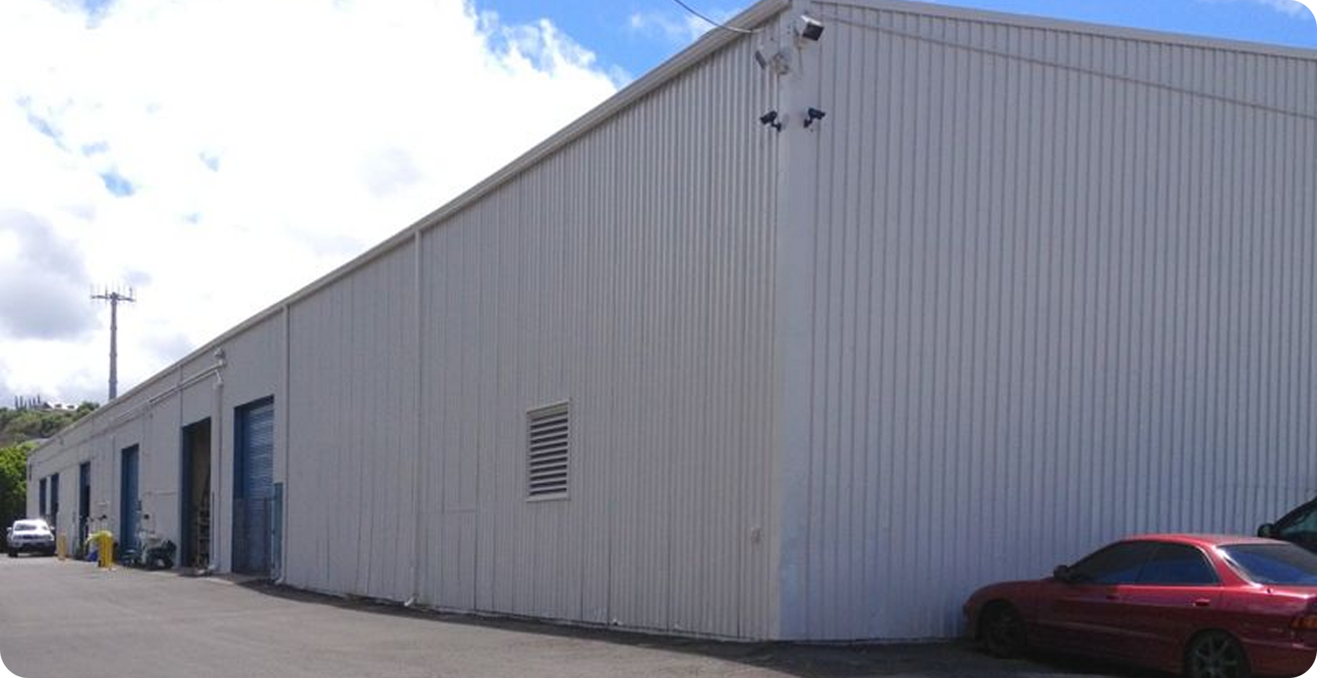 Large gray industrial building with blue doors, parked red car, and bright sky