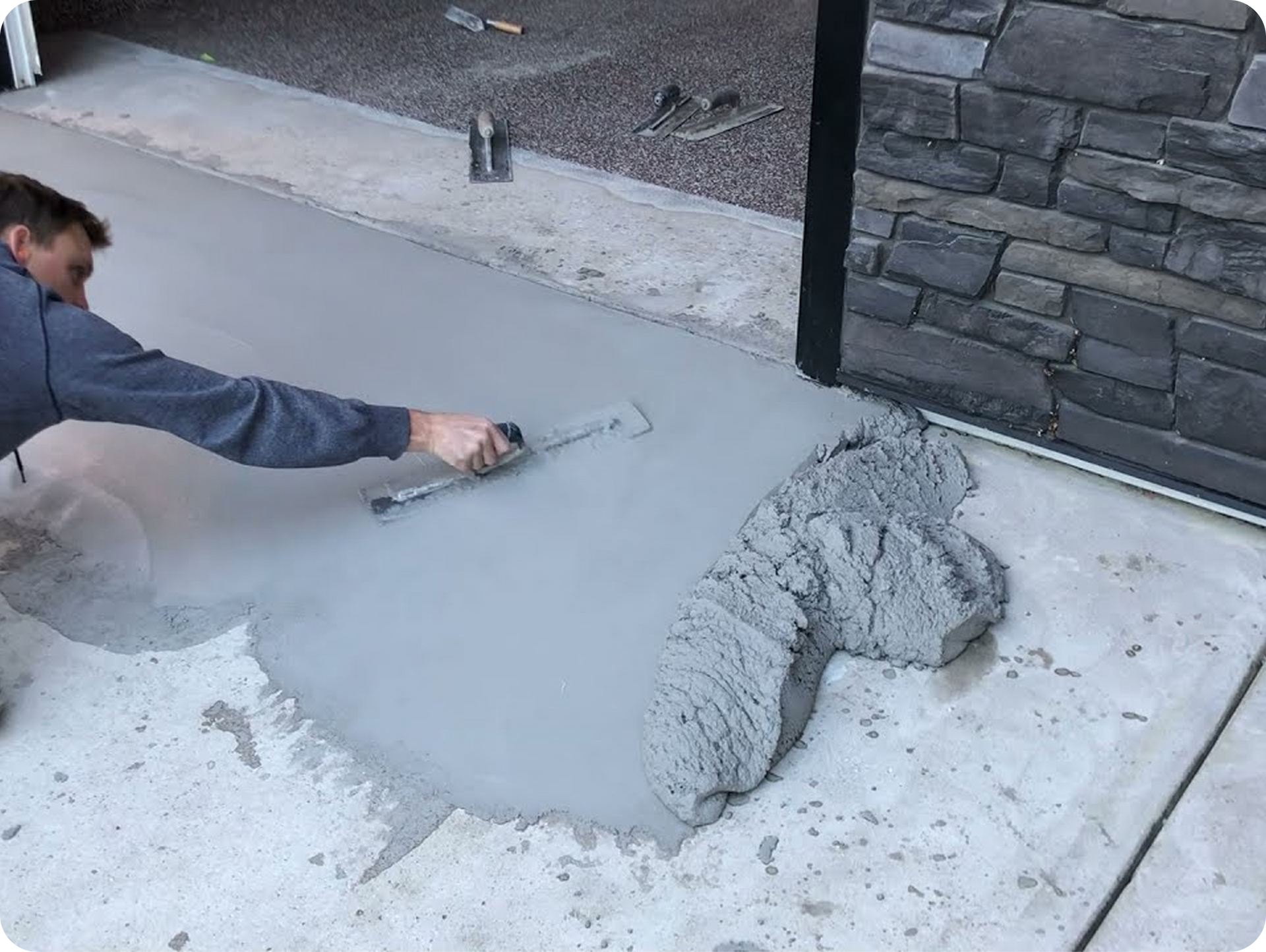Person smoothing wet concrete on a sidewalk beside a stone wall and house foundation