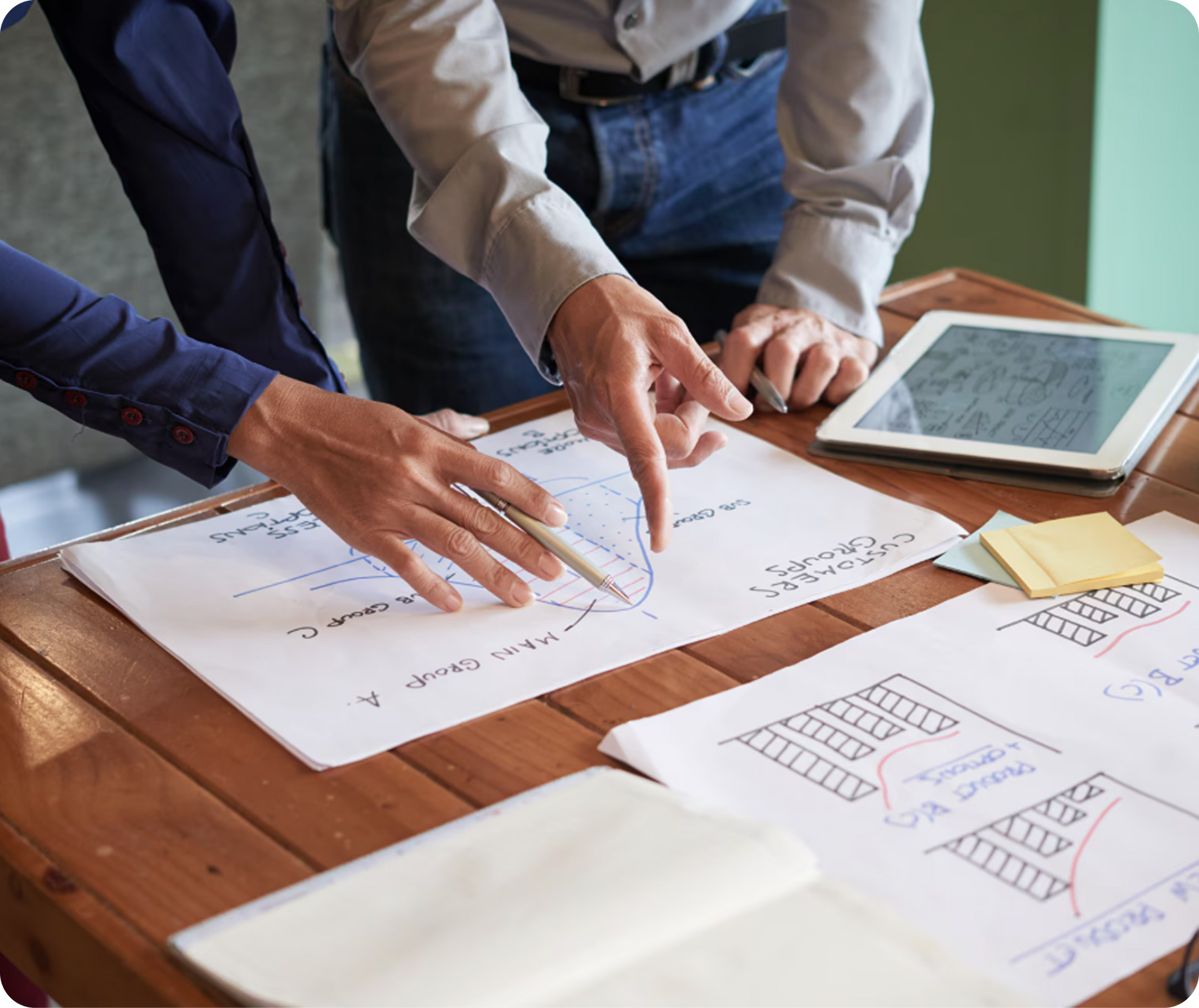 Hands reviewing paper wireframes on a wooden table beside a tablet and sticky notes.