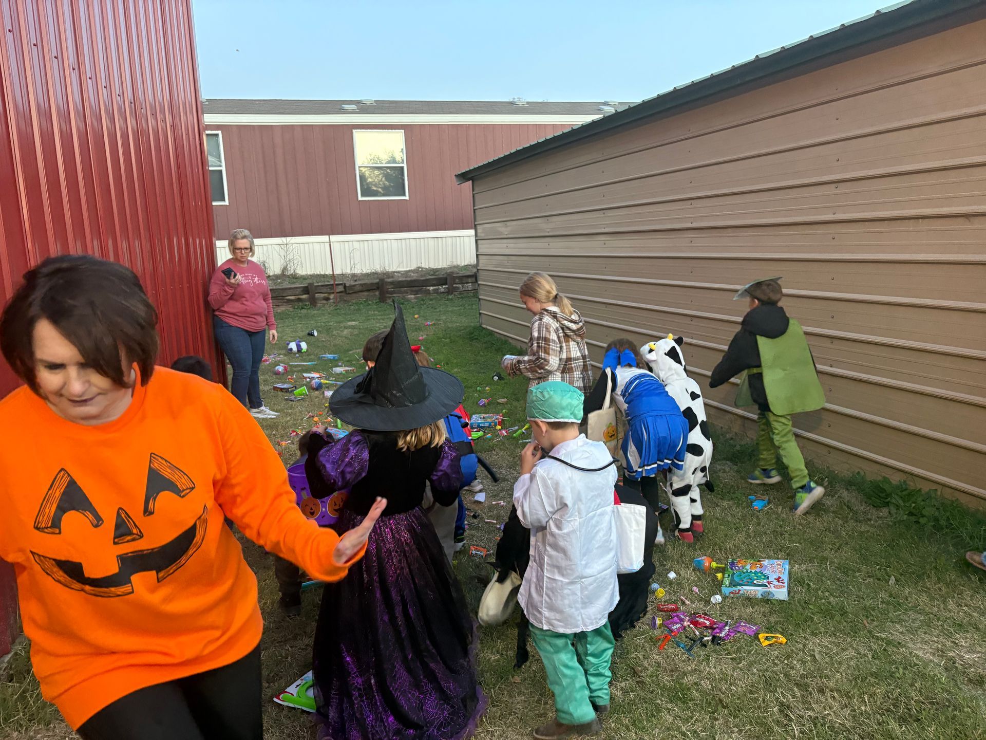 Children in Halloween costumes collecting candy outdoors, near buildings. Woman in orange shirt.