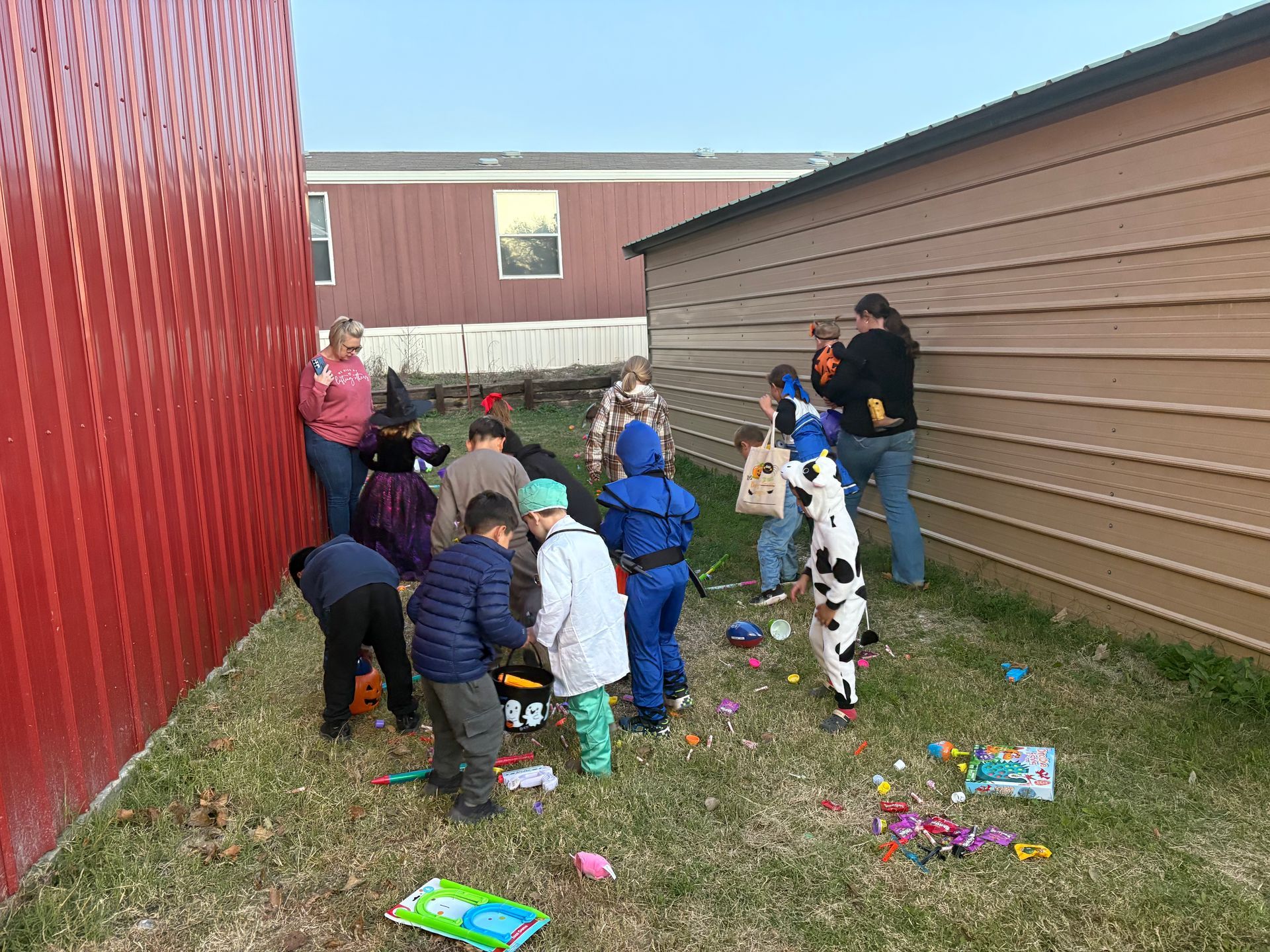 Children in costumes gather outdoors with adults. Toys are scattered on the grass between two buildings.