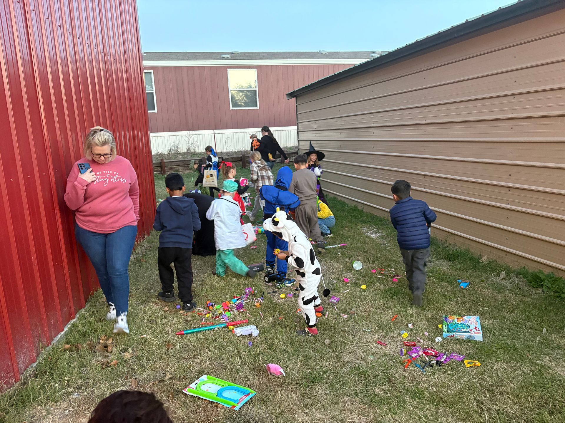 Children in costumes collecting colorful eggs in a grassy area, watched by an adult.