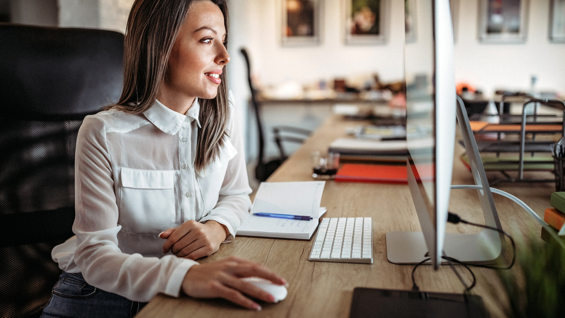 A person in a white collared shirt smiles while using a computer mouse at a desk in a bright office.
