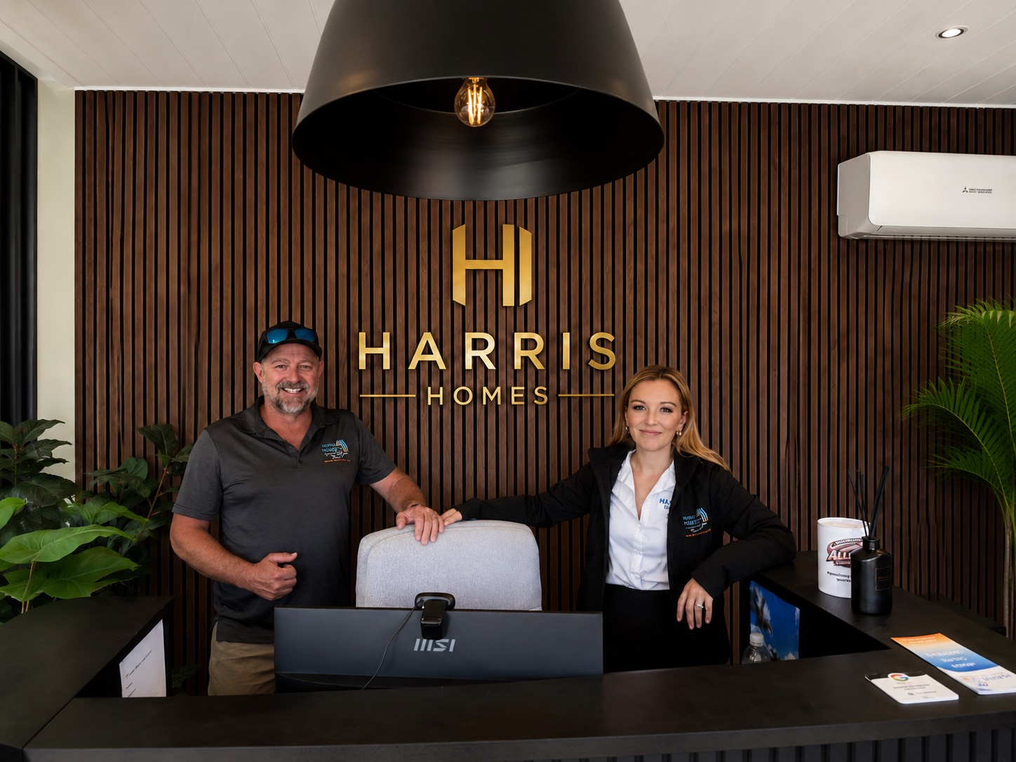 Two people behind a reception desk in a modern lobby with a Harris Homes logo on the wall. - Harris Design & Building Concepts In Jubilee Pocket, QLD