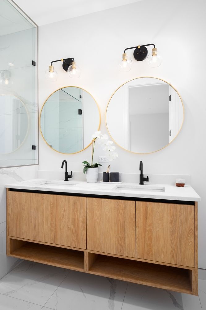 Modern bathroom vanity with two round mirrors, black sconces, and a wood cabinet beneath a white countertop. Harris Design & Building Concepts In Jubilee Pocket, QLD