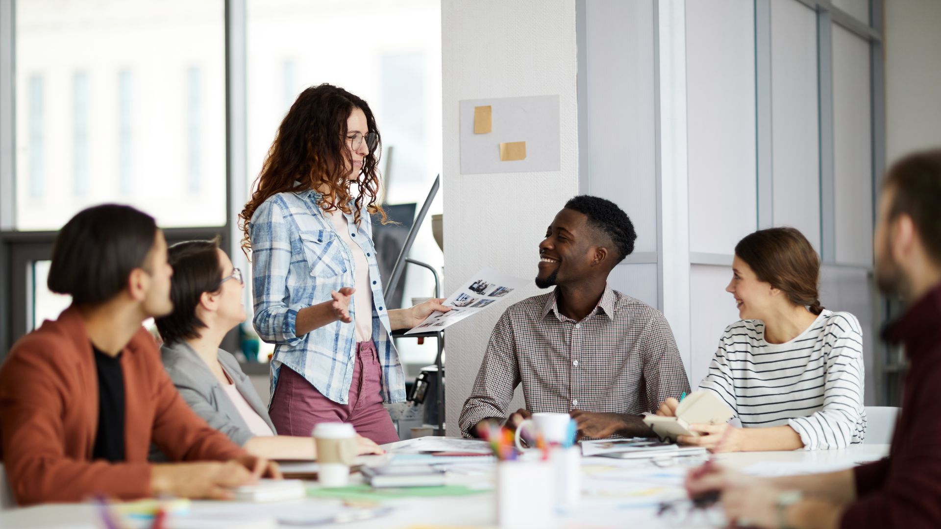 Group of people in an office meeting; woman presenting while others listen at a table with papers.
