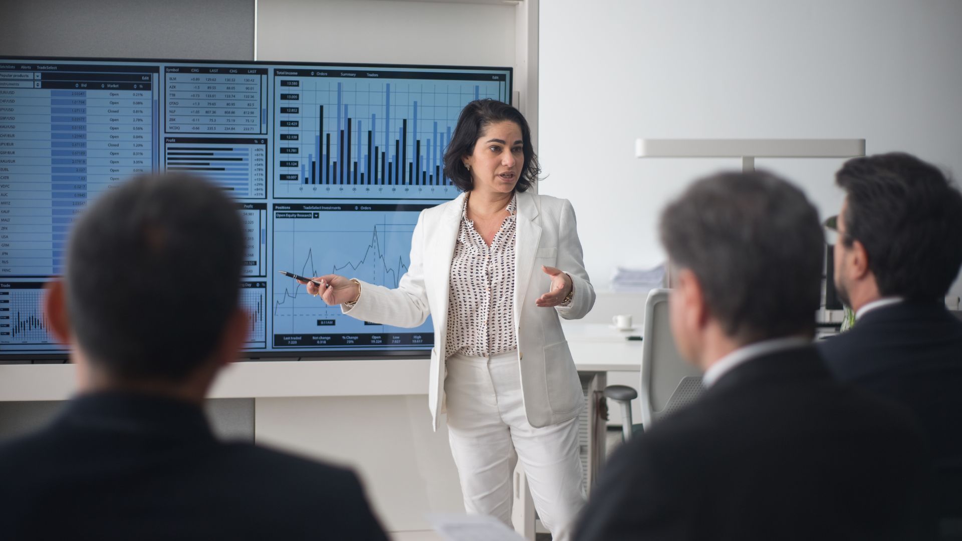Woman in white suit presenting data to colleagues; screens with graphs in background.