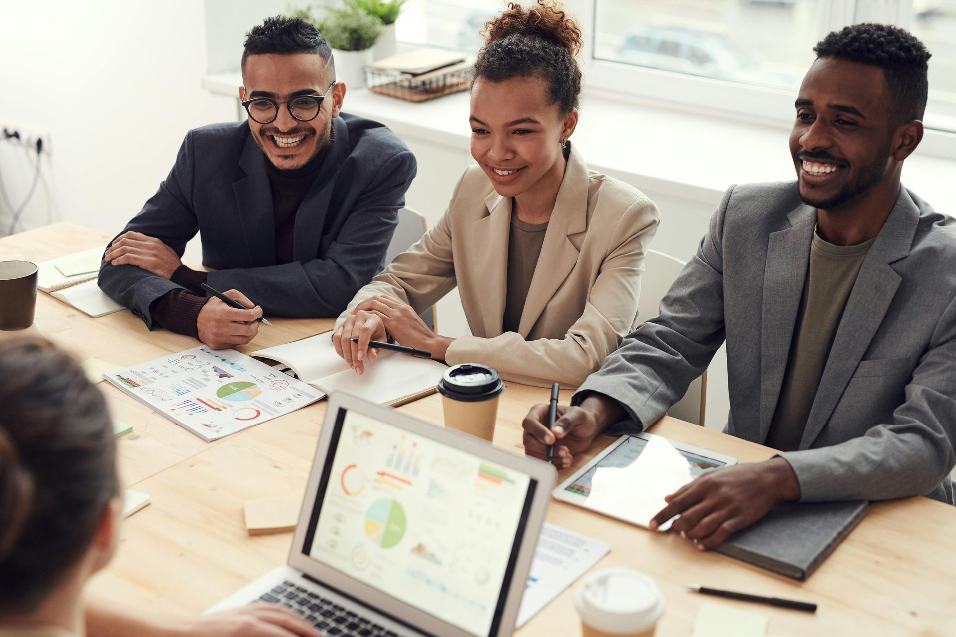 Three people smiling around a table with a laptop, documents, and coffee cups in an office setting.