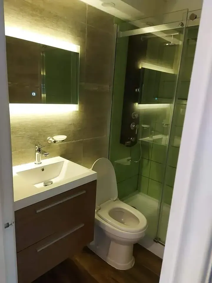 Baño moderno con espejo iluminado, lavabo con mueble, inodoro y ducha con mampara de cristal en azulejos verdes.