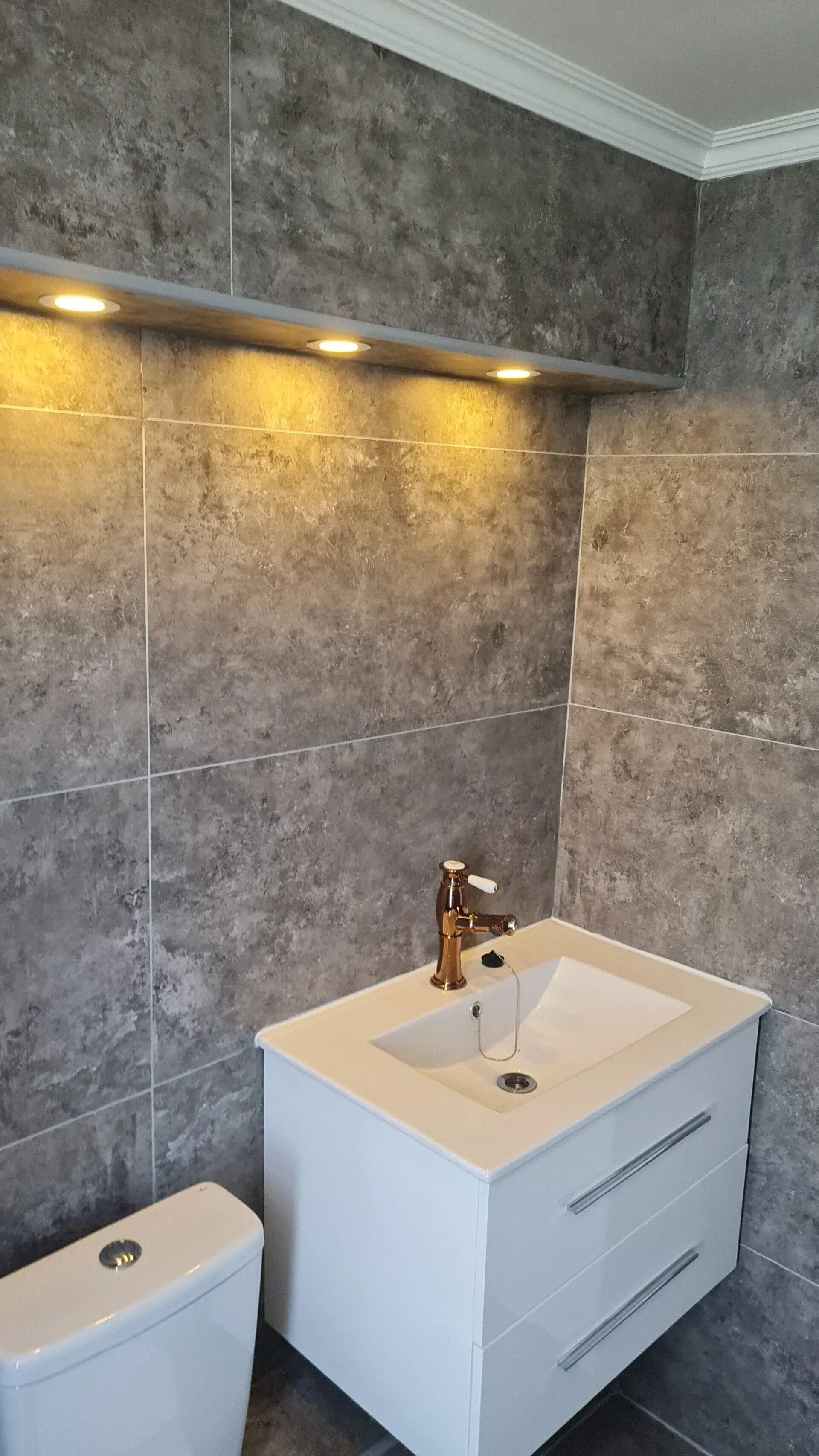 Baño moderno con paredes revestidas de azulejos grises, lavabo con tocador, inodoro e iluminación cálida bajo los gabinetes.