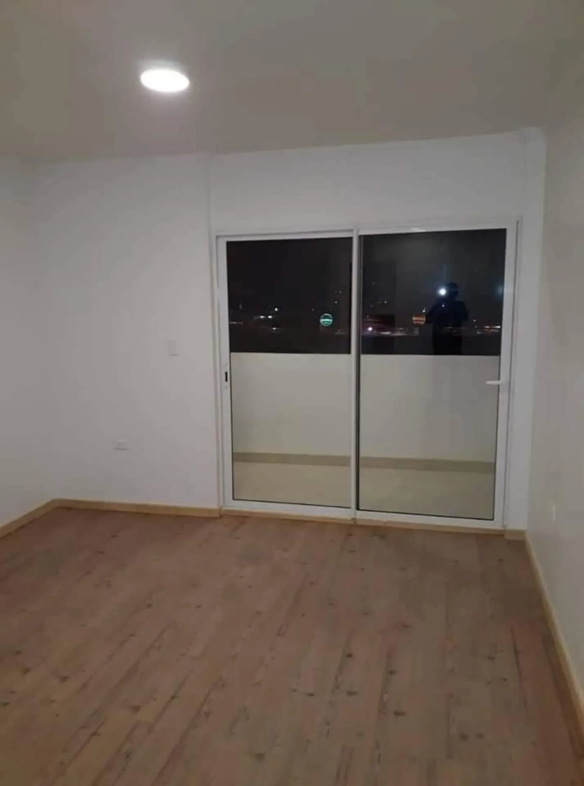 Habitación vacía con suelo de madera, paredes blancas, luz empotrada y puerta corredera de cristal que da a un balcón por la noche.