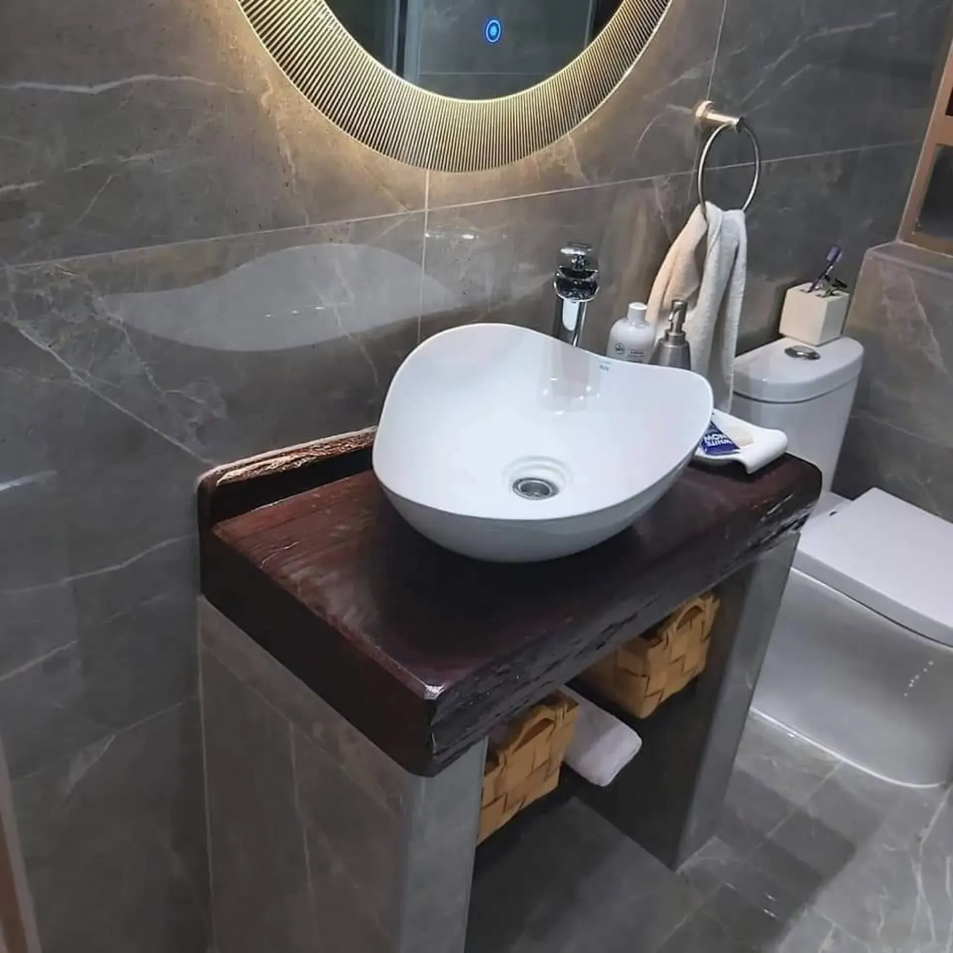 Baño moderno con lavabo sobre encimera de madera oscura, espejo de pared y paredes revestidas con azulejos de estilo mármol gris.