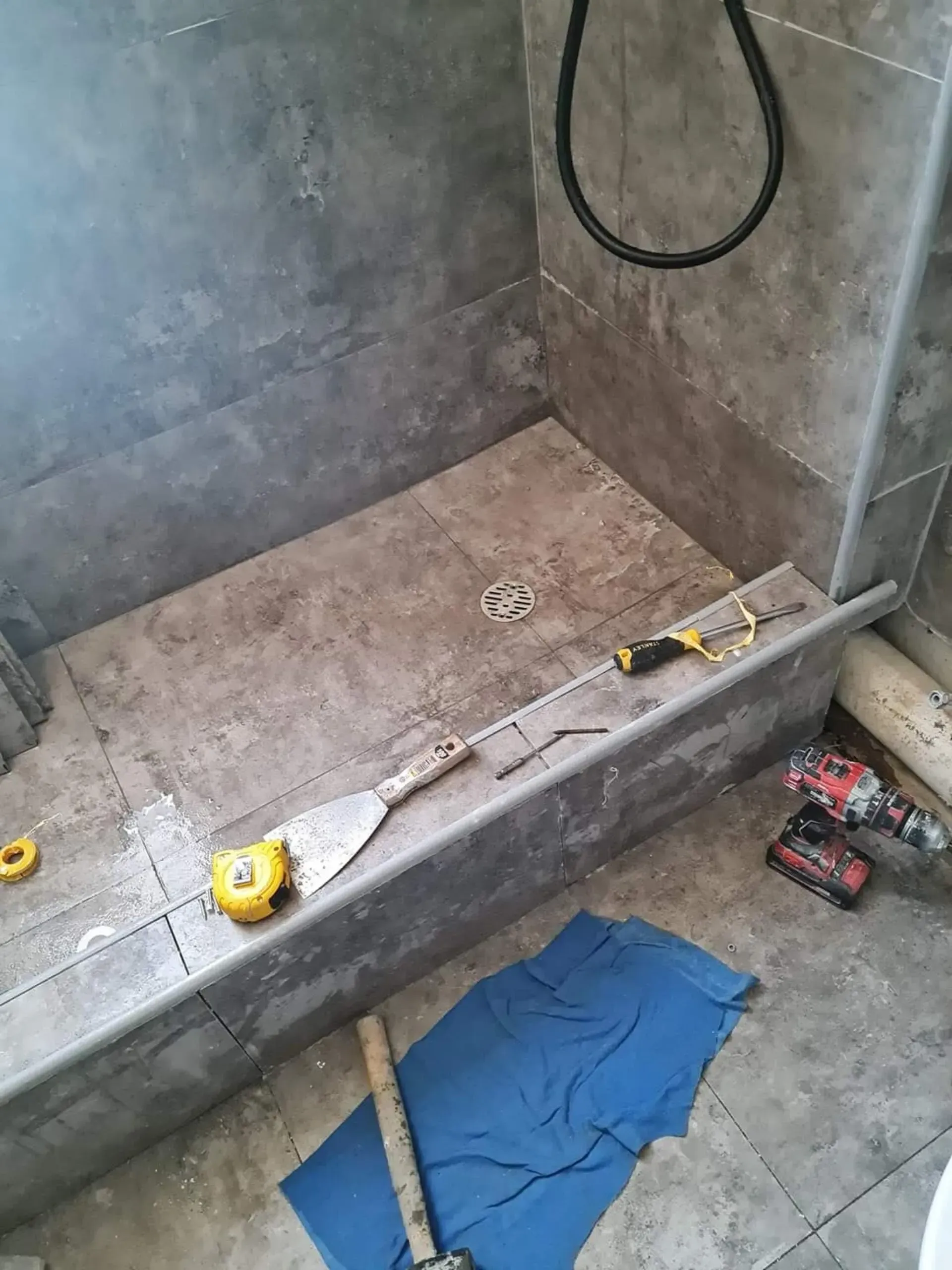 Zona de ducha del baño en construcción, con herramientas, una lona azul y una manguera en el suelo.