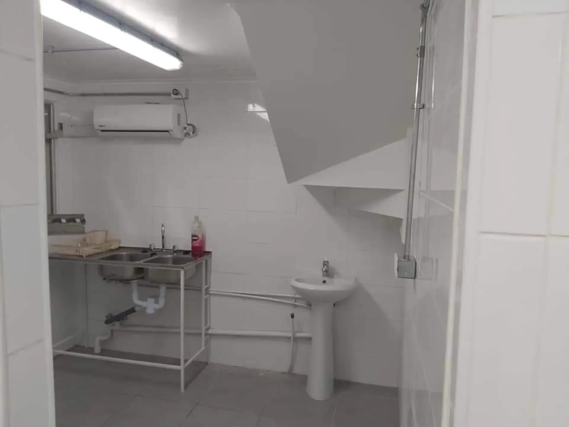 Pequeño cuarto de servicio blanco con fregadero, mesa de trabajo de metal y luz fluorescente.