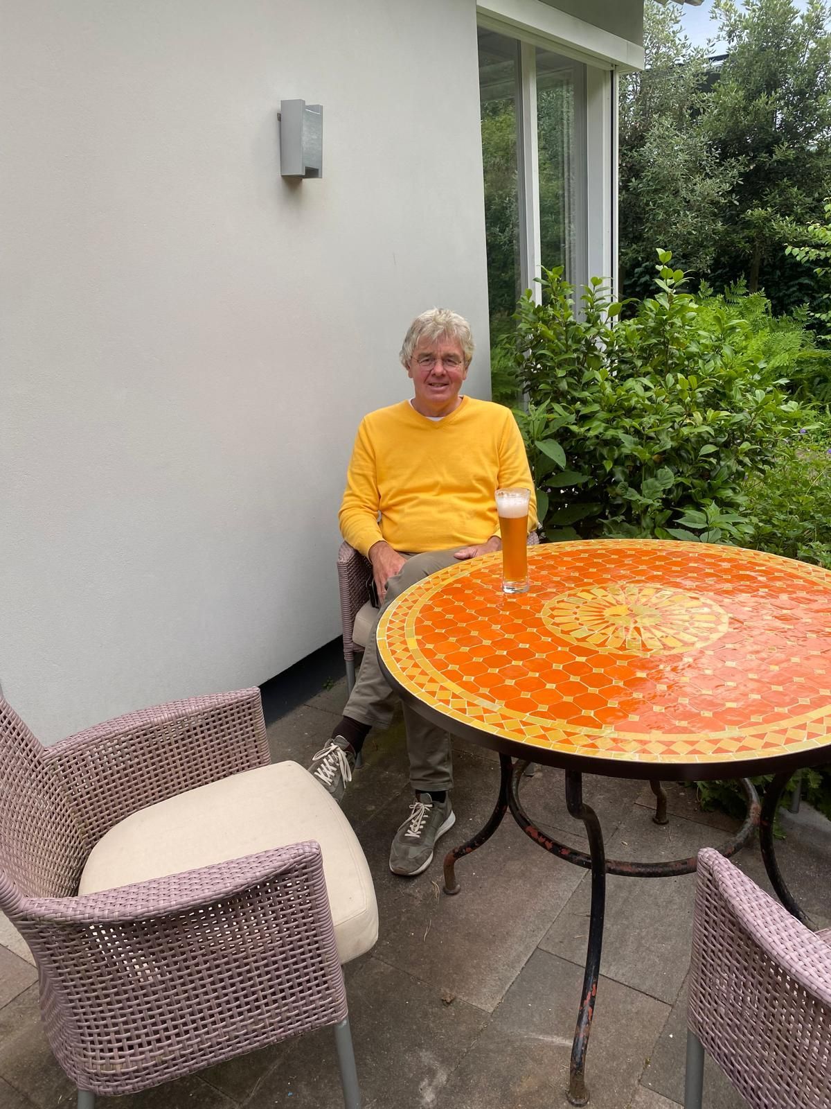Een man in een geel shirt zit met een biertje aan een oranje Marokkaanse mozaïektafel buiten, vlakbij een gebouw.