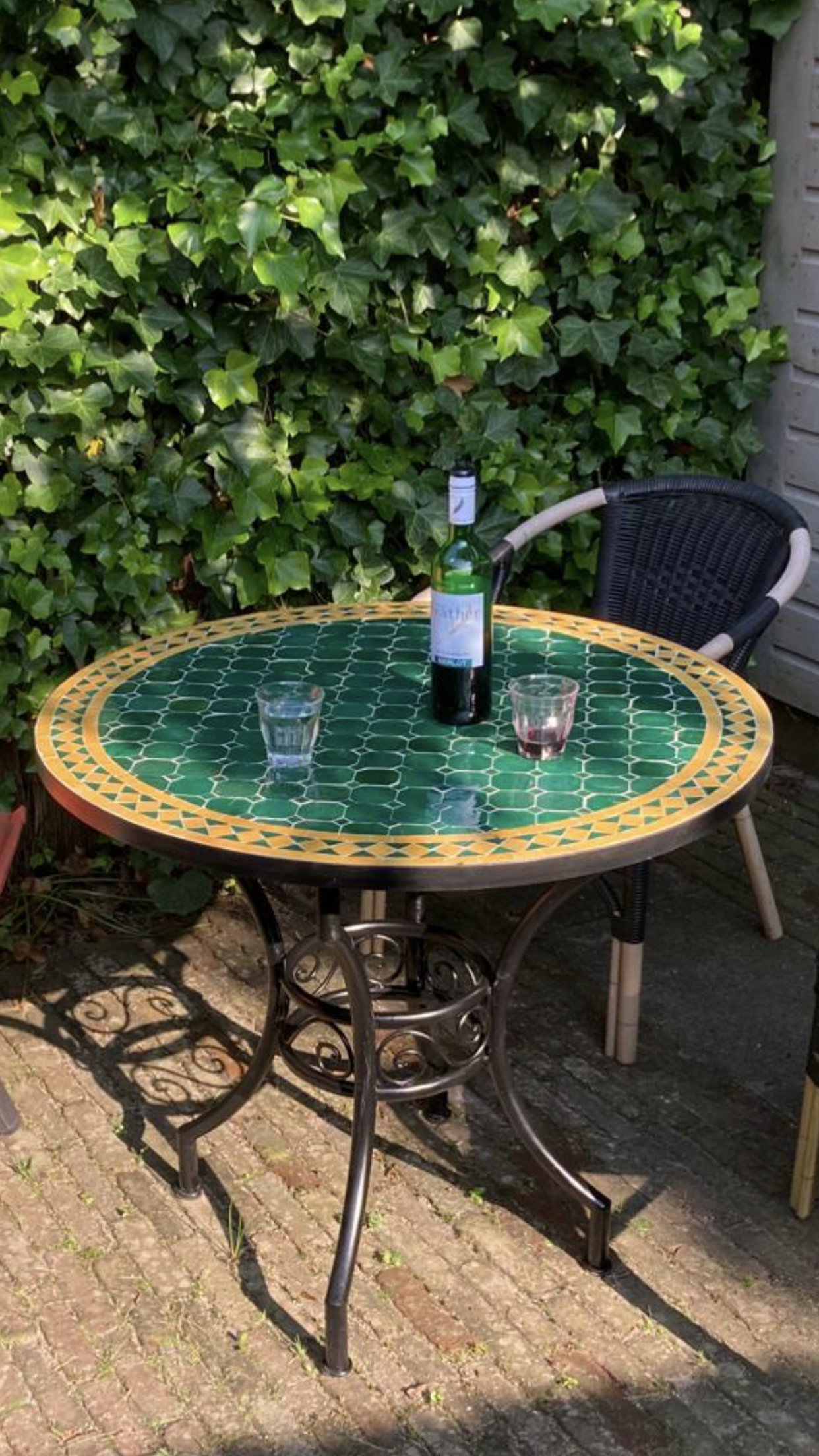 Een Marokkaanse mozaiek tafel met een fles wijn en glazen, op een terras, met een groene, lommerrijke muur erachter.