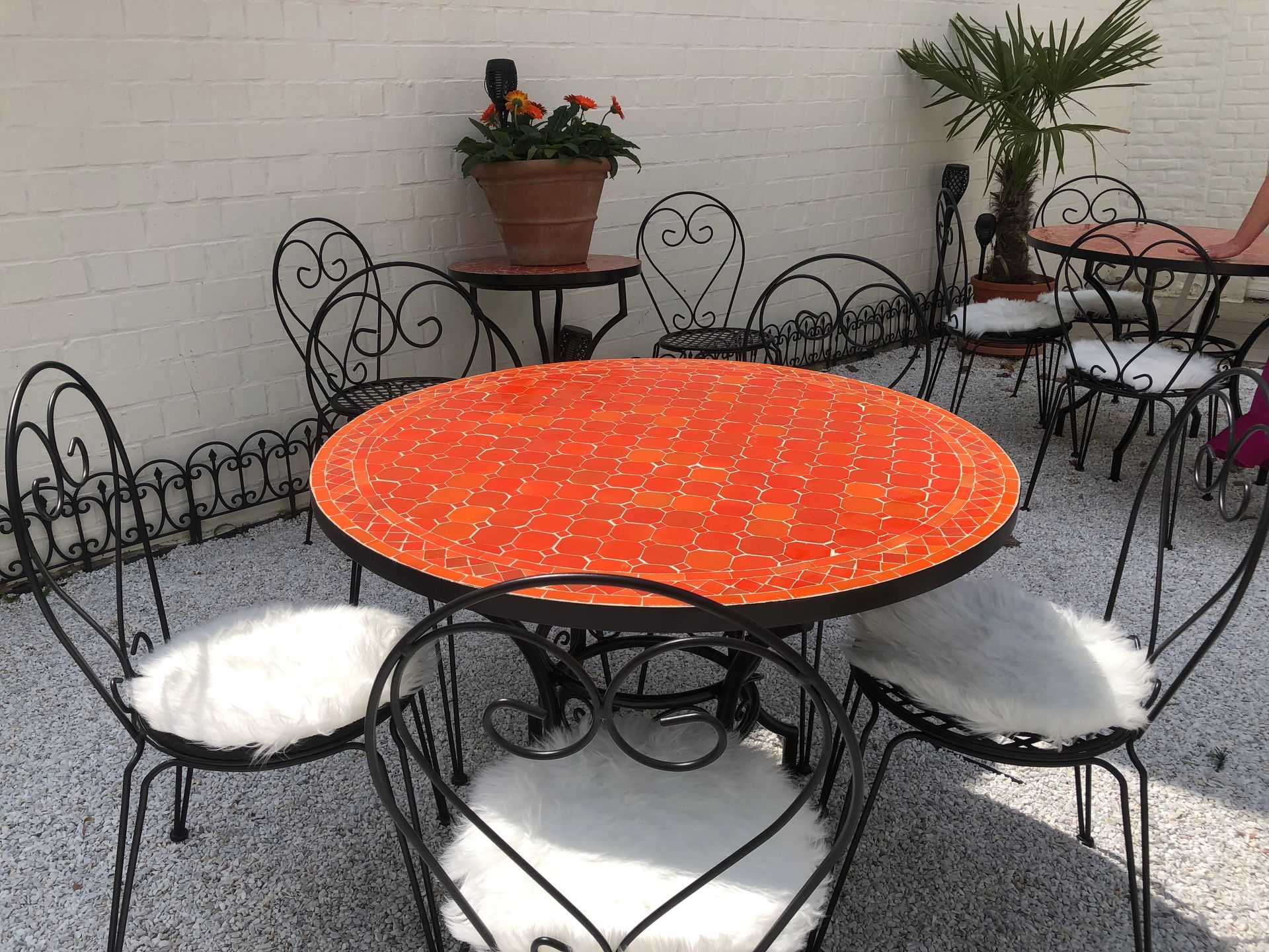 Ronde oranje Marokkaanse  mozaïektafel met sierlijke zwarte smeedijzeren stoelen op een zonnige binnenplaats.