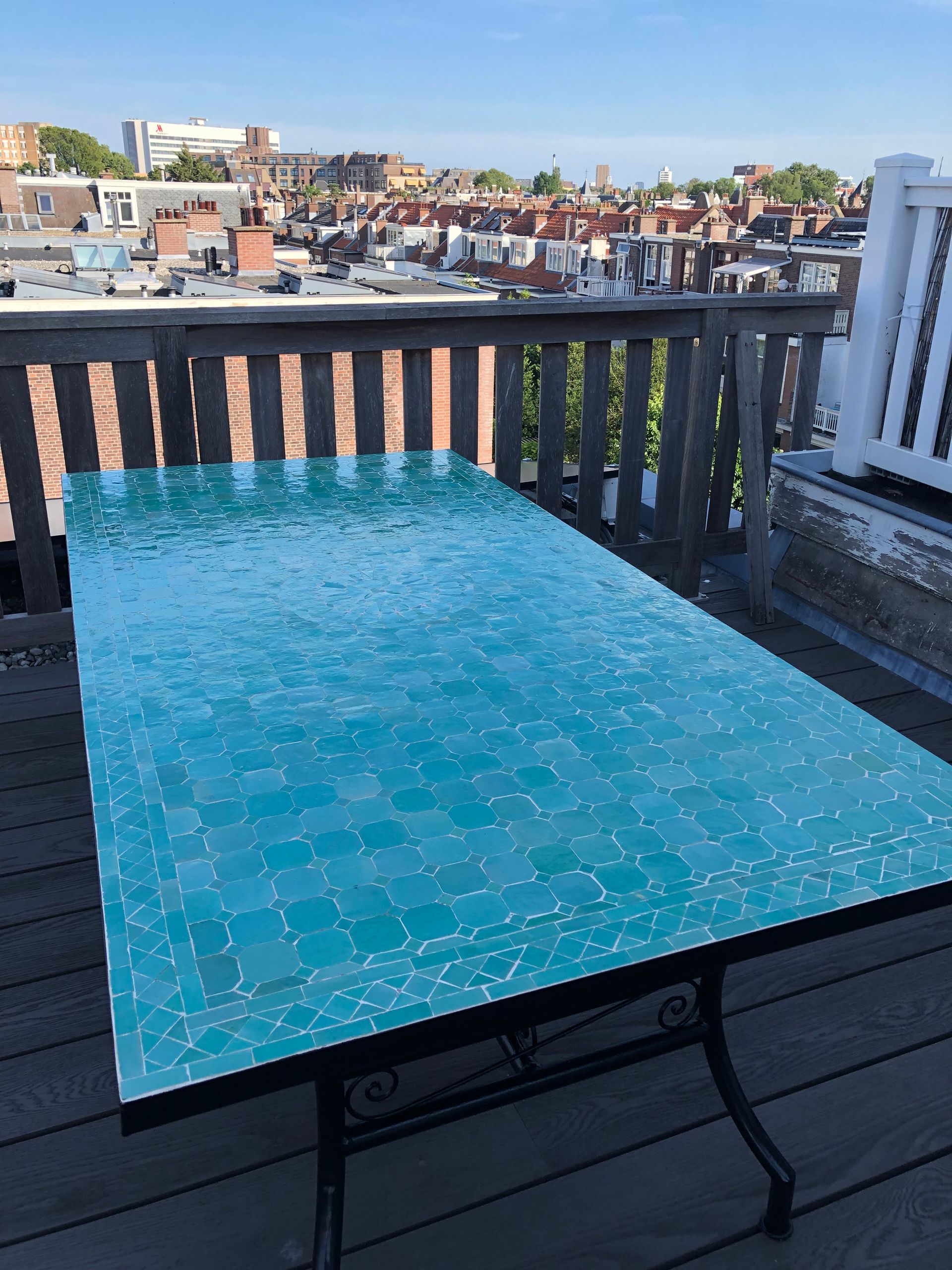 Een Marokkaanse mozaiektafel met turquoise tegels op een houten terras, met stadsgebouwen op de achtergrond.