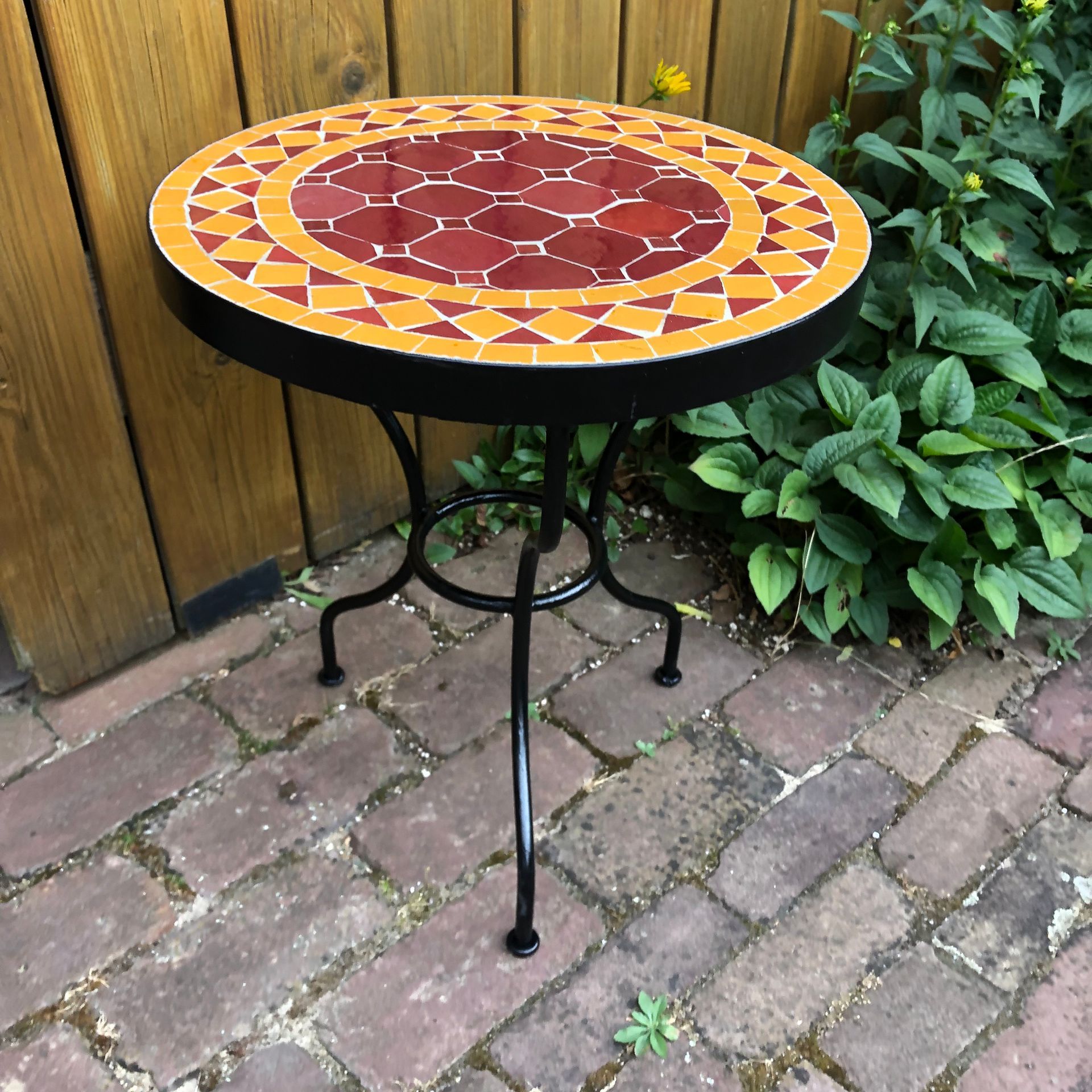 Ronde Marokkaanse mozaiek tafel met  zwarte metalen poten, geplaatst op een bakstenen terras naast een houten muur en groen.