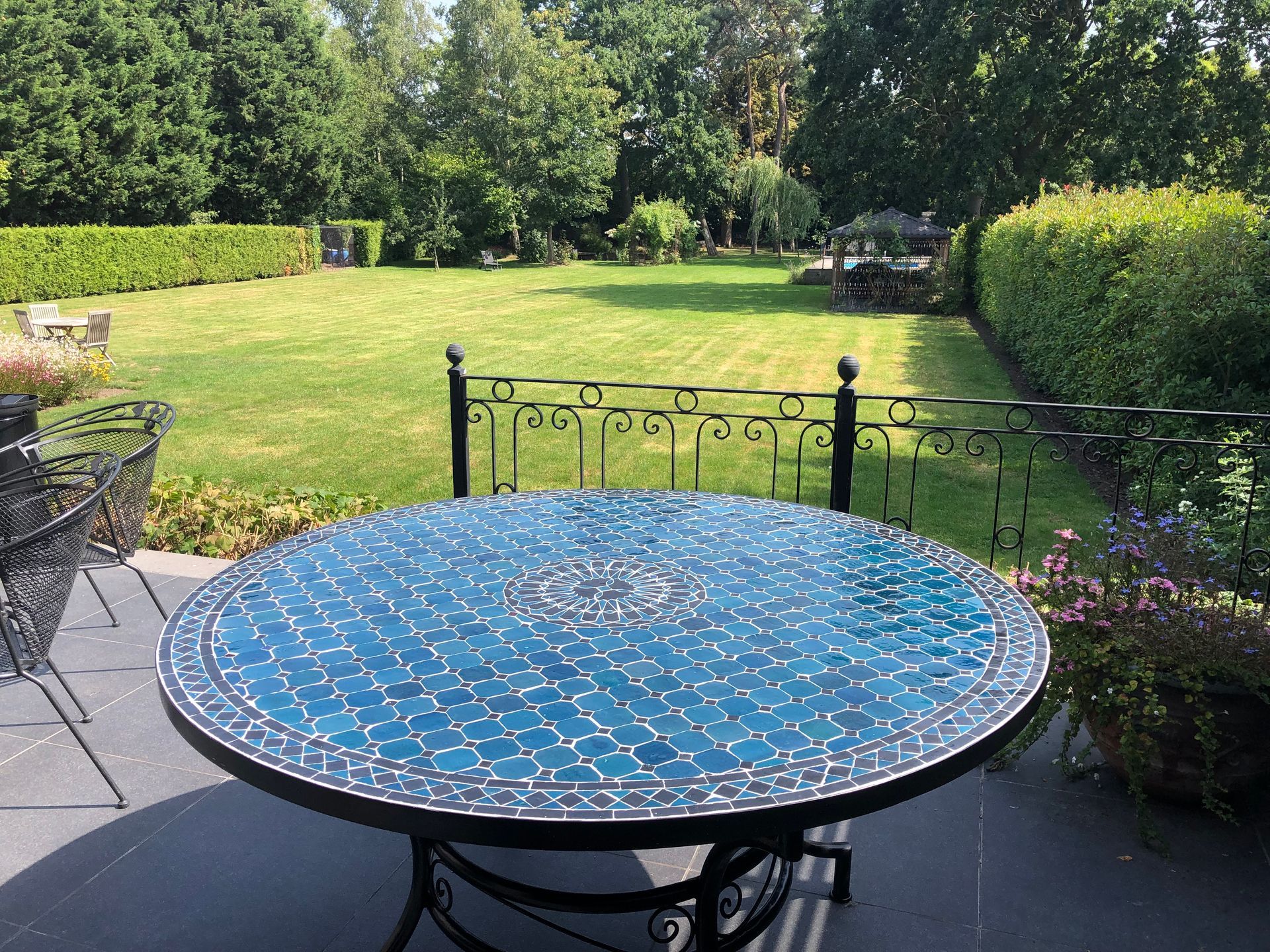 Tafel met mozaïekblad uit Marokko op een terras met uitzicht op een groen gazon en bomen.