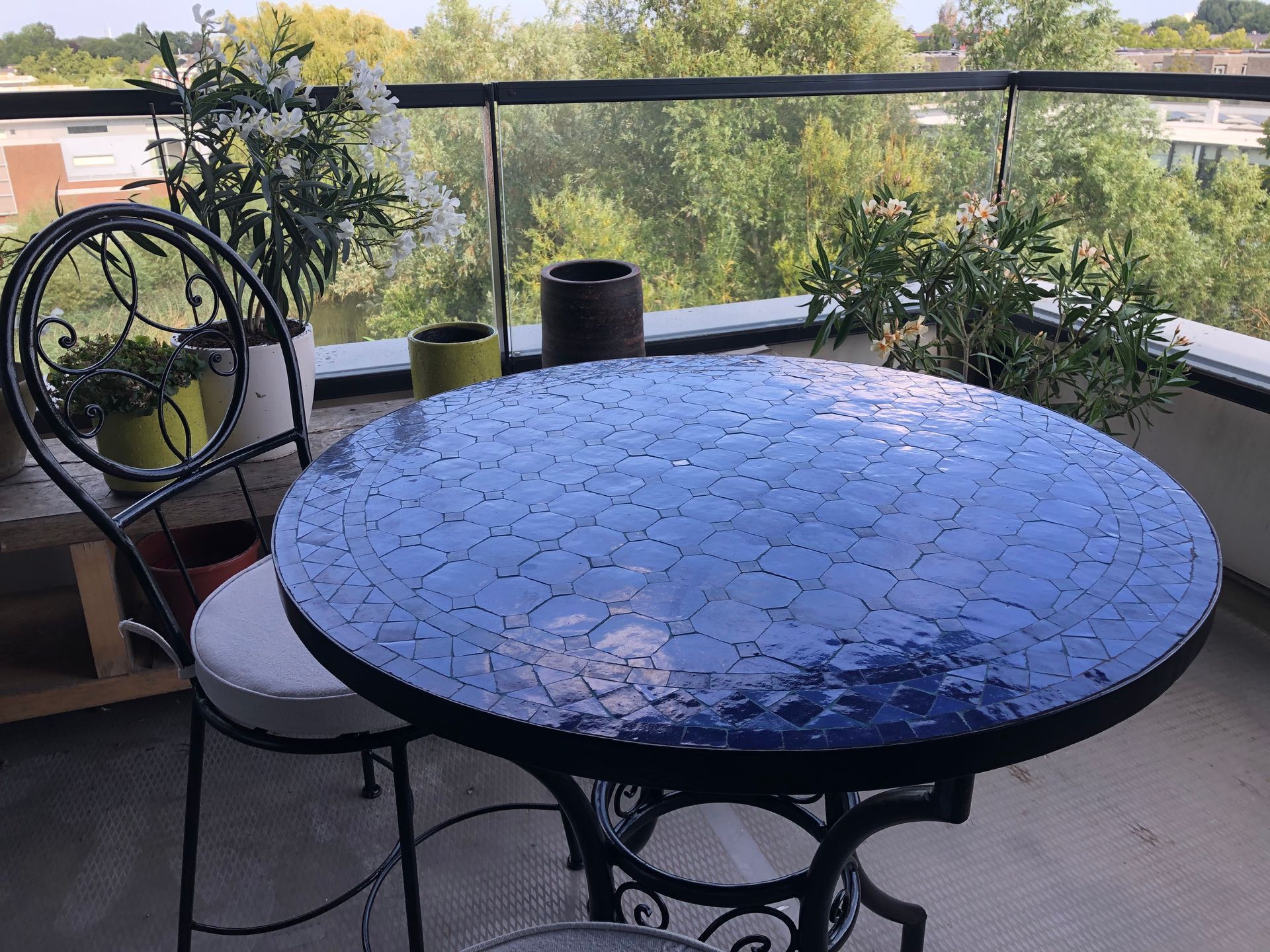 Balkon met Marokkaanse mozaïektafel, smeedijzeren stoel en potplanten. Bomen op de achtergrond.