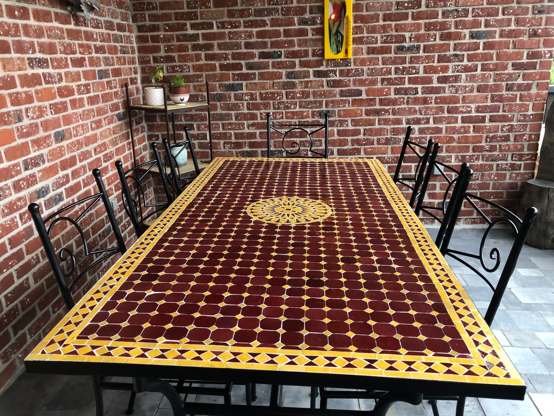 Terrastafel met een blad van rode en gele tegels en zwarte metalen stoelen, tegen een bakstenen muur.