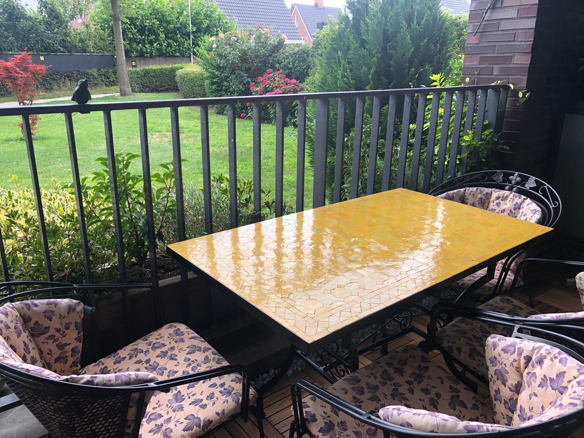 Een handgemaakte Marokkaanse  mozaiektafel met stoelen, met uitzicht op een grasveld, omgeven door een zwart metalen hek.