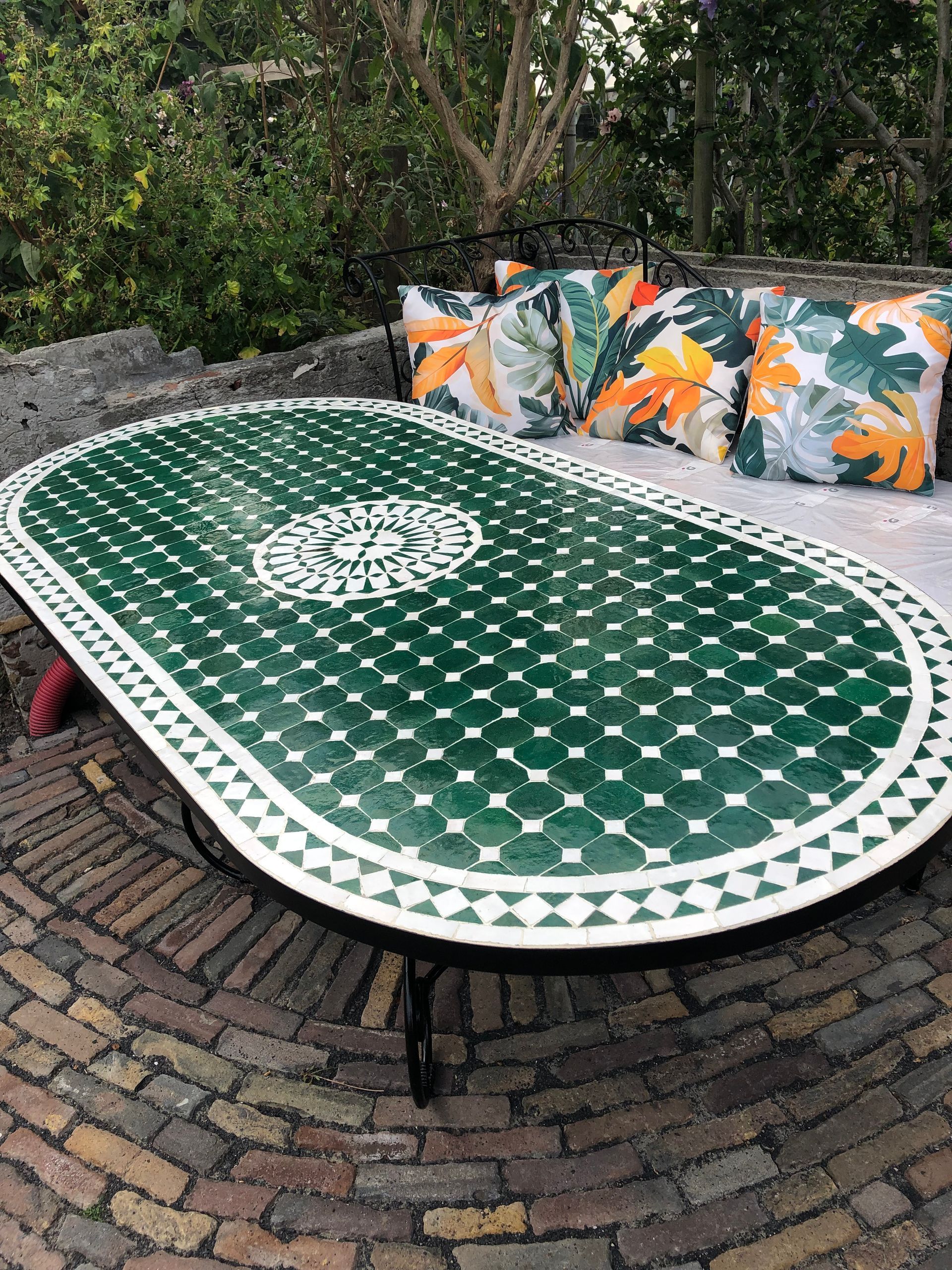 Ovale Marokkaanse mozaïektafel op een bakstenen terras met bloemenkussens op de achtergrond.