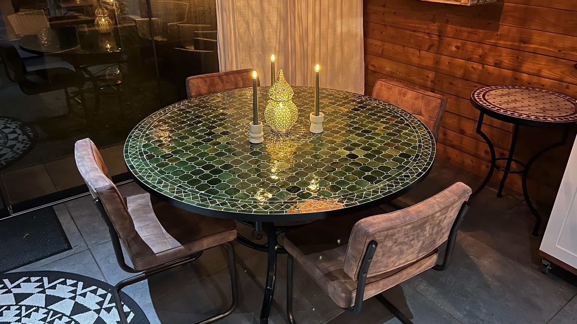Ronde Marokkaanse  mozaiektafel met vier stoelen en brandende kaarsen. Bakstenen muur en bijzettafel met mozaïek.