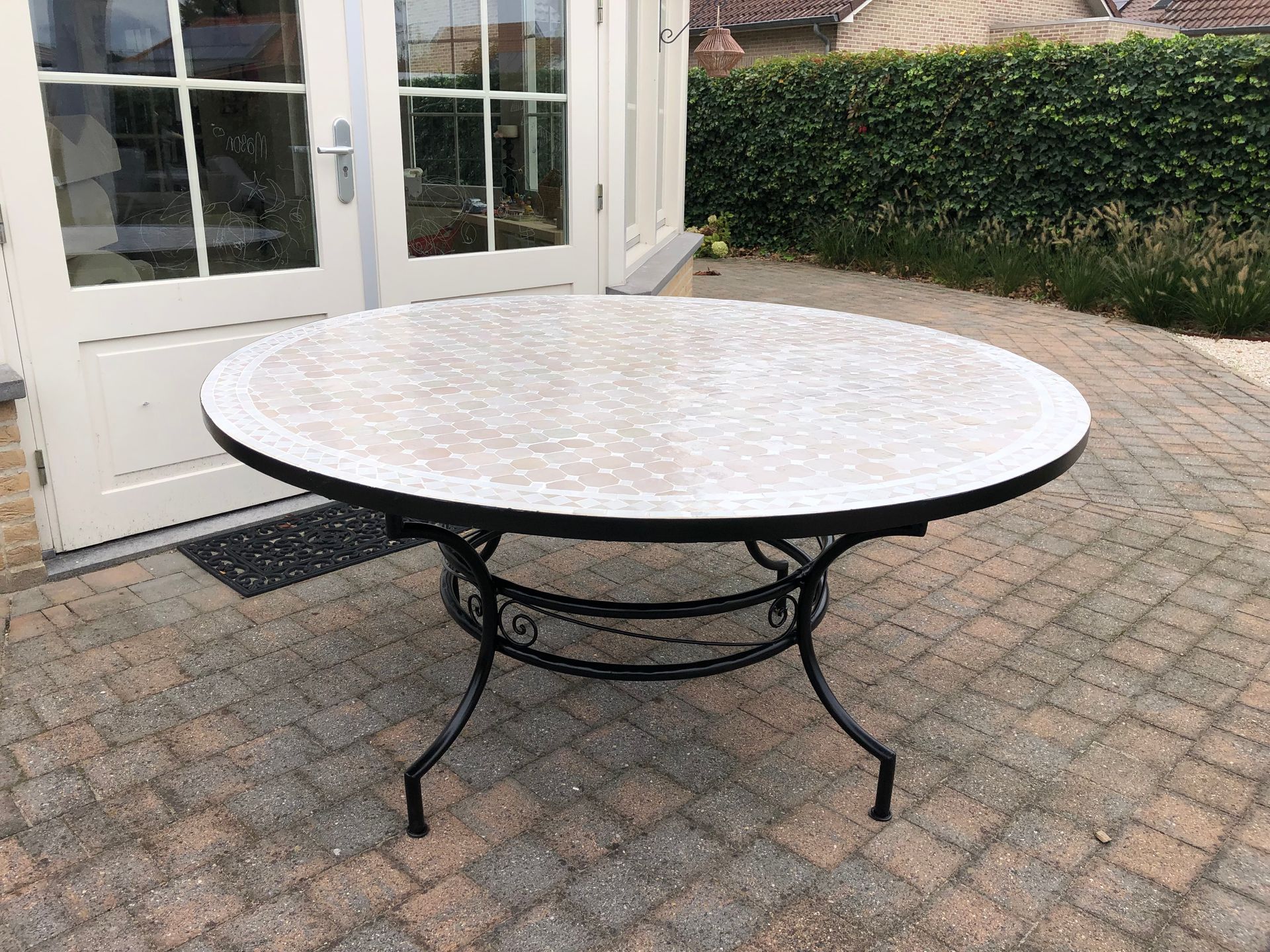 Ronde buitentafel met een mozaïekblad en een zwart metalen onderstel. Geplaatst op een stenen terras voor een glazen deur.