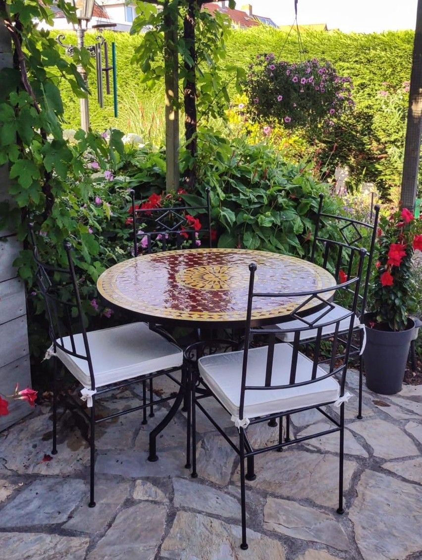 Terrasset met ronde Marokkaanse mozaiek tafel en vier smeedijzeren stoelen op een stenen terras, omgeven door tuin.